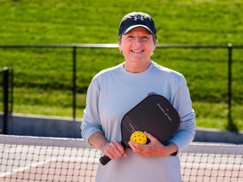 A woman smiling while holding a pickleball and paddle.